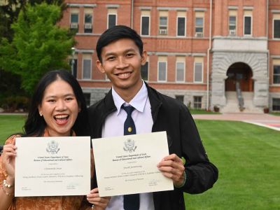 Two graduates proudly display their diploma certificates in front of a University Hall at UM