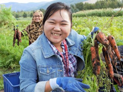 Two women harvesting carrots in a field, one smiling and holding a bunch of carrots, while the other holds up more in the background