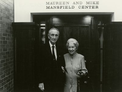 Maureen and Mike Mansfield standing together in front of the Mansfield Center. They are both smiling, and Maureen is holding Mike's arm.