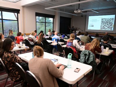 groups of people sit at long white tables looking at a projected image