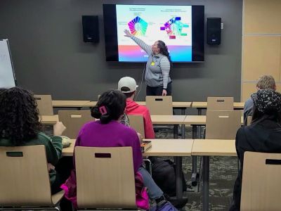 A role model explains a concept using a colorful visual slide to a room of high school students.