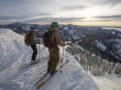 Two skiers pause on a snowy ridge at Snowbowl, looking out over a mountain valley at sunset