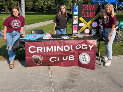 Crim Club leaders stand at an informational table with a chalk outline on the ground at the student Welcome event..