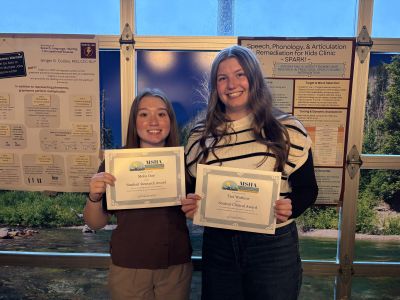 two students holding awards in front of posters. one award is for student research and the other is for student clinical.