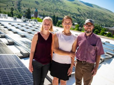 Eva, Maddie, and Paul smile on top of a sunny roof with solar panels in the background