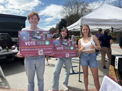 three students pose, two holding signs that share information about the Kless Fee increase campaign