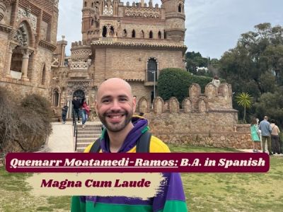 Quemarr stands in front of castle, grinning widely at camera