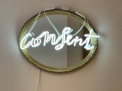 neon sign on mirror, neon is white and reads 'consent' in cursive, mirror has gold frame