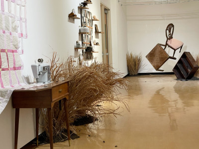 view of lefthand side of gallery including paper quilt, sewing machine, wood quilt, grasses, and hanging furniture in back