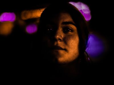 a girl partly in shadow looks upward at the camera, purple lights flaring behind her and reflected in her eyes, while an orange light glows on the right side of her face