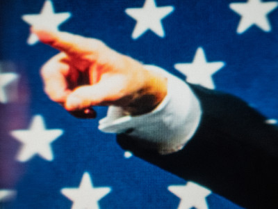 in front of the stars of the US Flag, a man's hand points straight up to the left