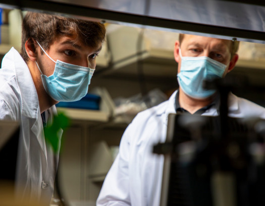 Two people wearing face masks and lab coats look at a computer monitor