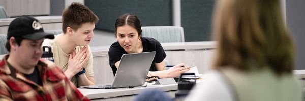 two students working together at a laptop with another student and the teacher in the foreground