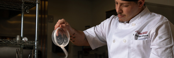 chef in a kitchen pouring spices from a bowl into another container not shown in the image