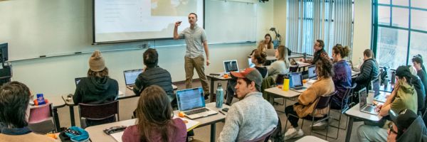 students seating at tables looking at teacher who is gesturing at image on a projection screen