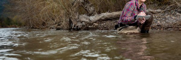 student crouching in a river taking notes