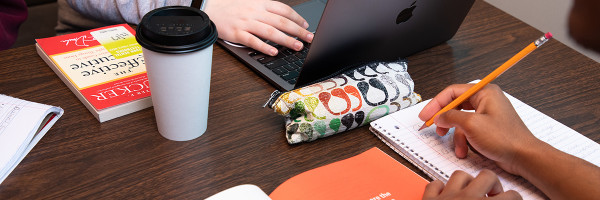 close up view of a table with two students studying one writing in a notebook and the other working on a laptop