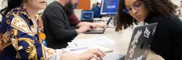 two students working together on a laptop with other students working on laptops in the background
