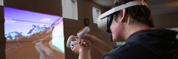 student wearing a virtual reality headset and holding controllers pointed at an image projected on a classroom wall