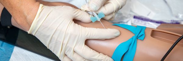 two gloved hands inserting a needle into the arm of a medical practice dummy