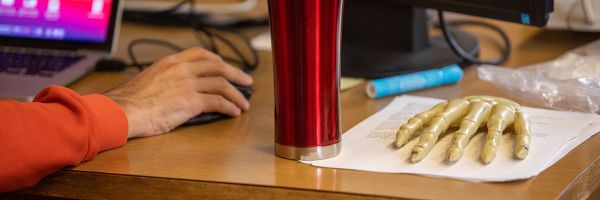 desk with a hand holding a mouse next to a model of the bones of a human hand resting on some papers