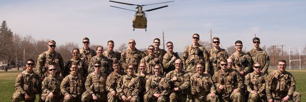 two rows of rotc students posing for the camera with a chinook helicopter hovering in the background