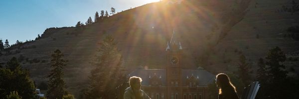 two students sitting on the UM oval talking with university hall in the background and the sun dramatically shining from behind mount sentinel