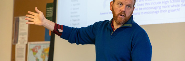 professor in front of a class speaking while holding an open book with a screen projected behind