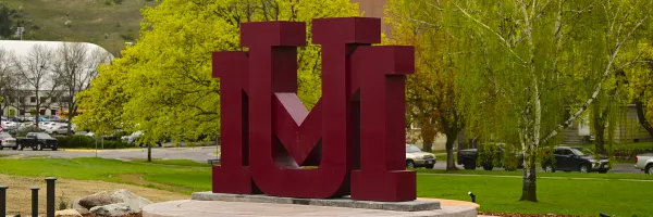Maroon intertwined block letter "U" and "M" sculpture sign on a concrete base near the Madison street bridge close to the University of Montana campus in Missoula, MT with grass and trees in the background.