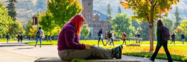 A student sits on a bench on the Oval as other students walk across campus