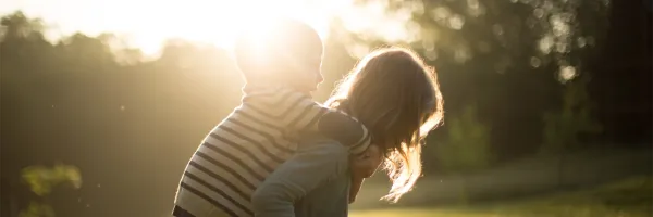 Person carrying a child on their back outdoors, backlit by warm sunlight with trees softly blurred in the background.