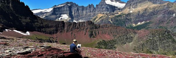 person sitting and reading a book on a rocky hill next to tall mountains with patchy snow