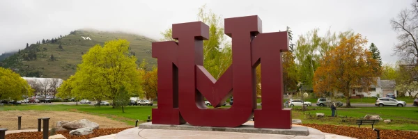 Large Maroon University of Montana block "UM" logo sculpture, with trees and Mount Sentinel’s white "M" visible on a misty mountainside behind.