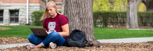 student working on a laptop sitting outside leaning against a tree
