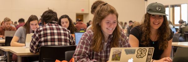 two students working on their laptops and smiling with many other students working at other tables in the background