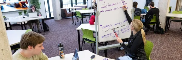 student drawing equations on a white board with tables of other studying students in the background