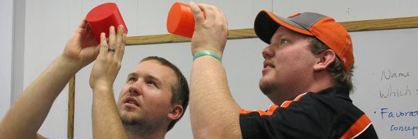 two math teachers in front of a white board holding up and looking at orange shapes