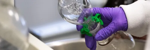 A UM doctoral candidate conducting research in a lab on the �Թ��� campus with a gloved hand holding laboratory glassware with a green clamp over a lab sink..