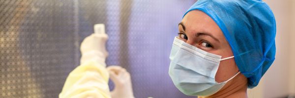 person in mask and other protective gear looking at camera while working with a pipette and vial in the background