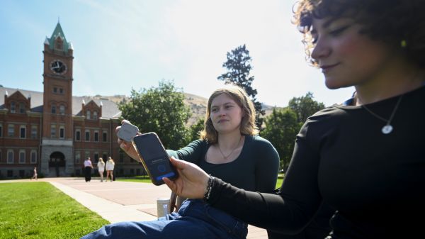 Two female students are seen on the Oval, one holding a Brick device, and the other tapping her phone to the Brick device to take intentional breaks from her phone.
