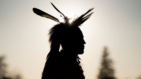 Silhouette of a dancer in regalia showing a head roach and feathers with the sun glistening behind