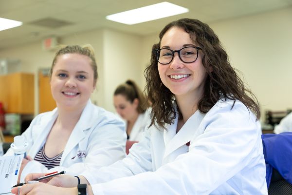 Two pharmacy students in lab coats smiling from their classroom seats