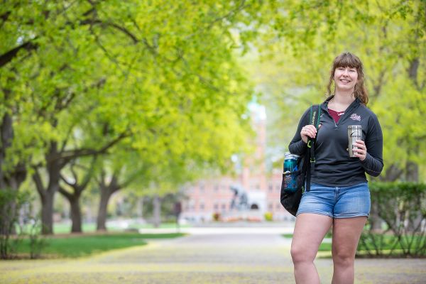 Female student holds backpack and Montana coffee mug in front of trees, the griz statue, and Main Hall