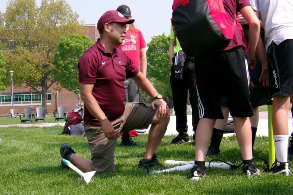 A role model kneels in grass as they demonstrate a rocket-launching activity for a group of middle school students.