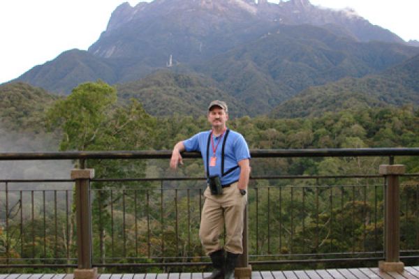 Tom Martin posing along a fence in front of a steep tree covered mountain