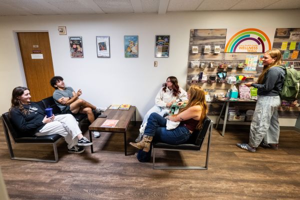 Six students stand together talking and laughing near the Wellness Wall inside the CHC Wellbeing/SARC office. The group appears relaxed and engaged in conversation, gathered in the welcoming space around the wall display.
