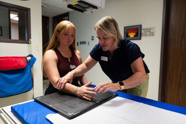 A medical provider positions a student’s arm for an X-ray, guiding them to ensure proper alignment while the imaging equipment is prepared.