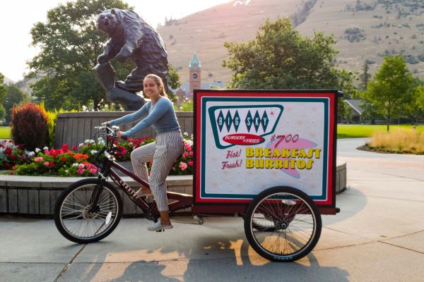 A girl smiles while posing in front of the  griz statue on dining's cargo bike