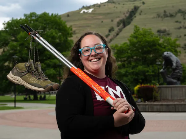 Brittany smiles for a portrait while holding hiking poles with a pair of hiking boots dangling from their shoelaces from the hiking poles