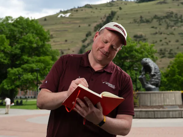Colin, wearing University of Montana apparel, stands and writes in a notebook